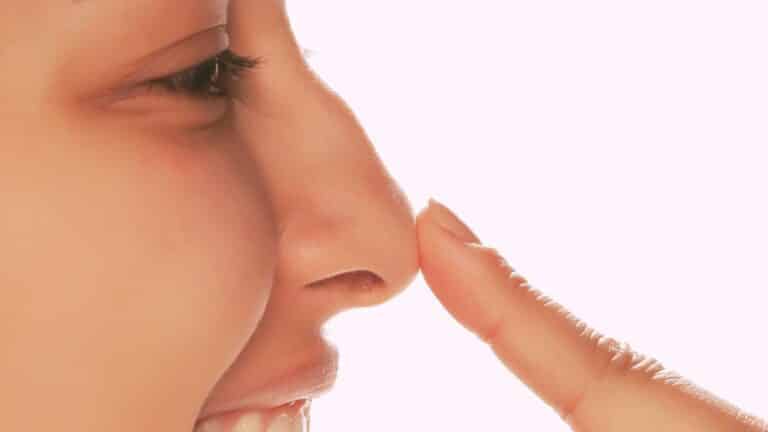 Nase ständig verstopft: 5 Anzeichen, die du nicht der Nase zuschreibst. Lachende Frau fasst sich mit dem Finger an die Nase.